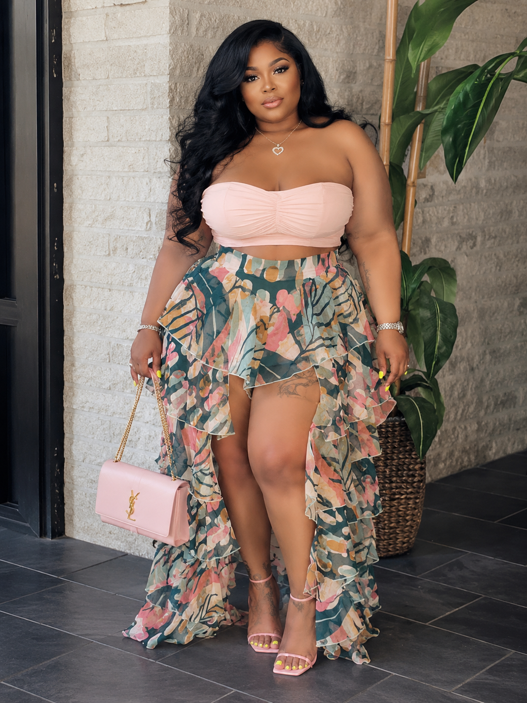 Woman in a floral dress and pink top standing against a stone wall.